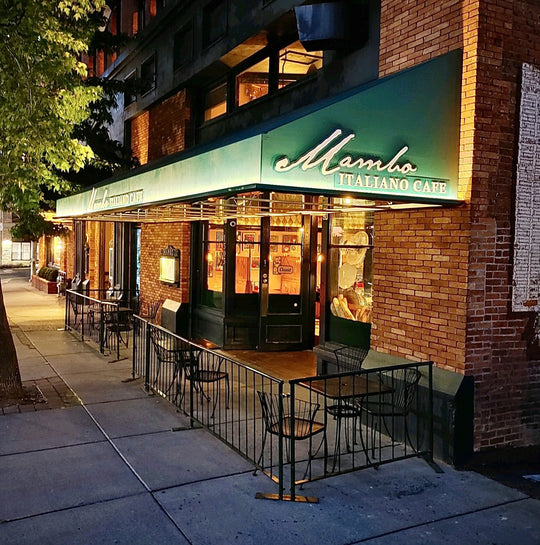 View of the Mambo storefront from the side. Green awning with logo, warm light coming from windows, patio tables out front.