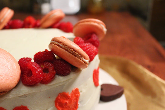 2022 Valentine's cake. White frosted red velvet cake with red hearts piped around the edge, chocolate hearts on the plate, and sliced raspberries and light pink macarons on top. 