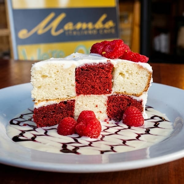 Checkered red velvet and vanilla cake slice with raspberries, cream, and chocolate drizzle on a white plate.