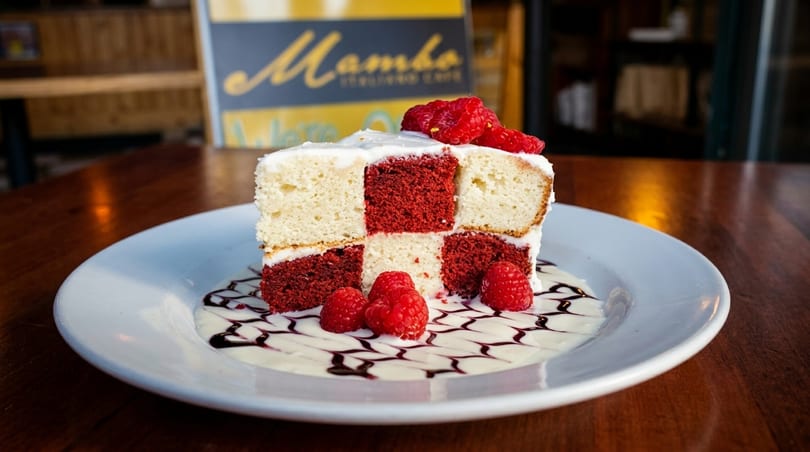 Checkered red velvet and vanilla cake slice with raspberries, cream, and chocolate drizzle on a white plate.