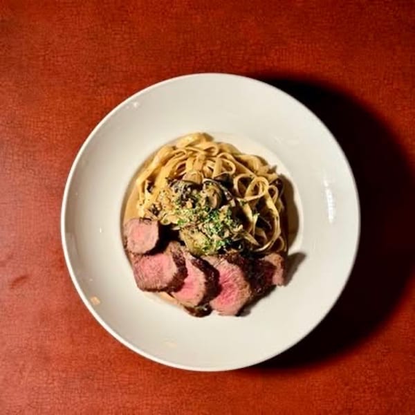 Sliced steak with creamy mushroom pasta, garnished with herbs on a white plate.
