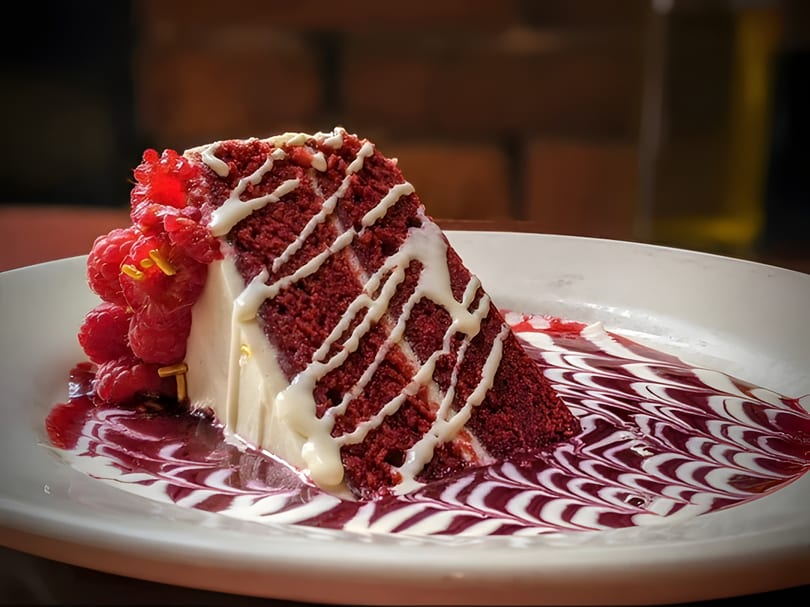 A slice of red velvet cake with cream cheese frosting, raspberries, and berry sauce.