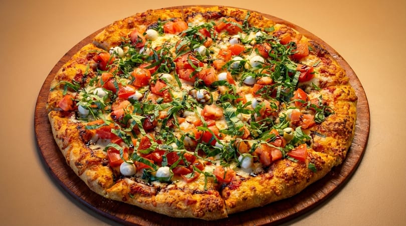 A whole pizza with diced tomatoes, mozzarella balls, and fresh basil on a wooden board.