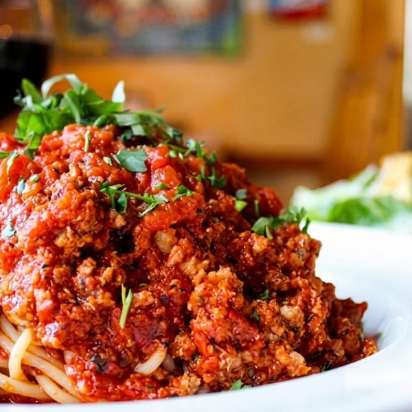 Spaghetti with meat sauce and fresh herbs, with a glass of red wine and salad in the background.