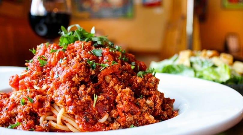 Spaghetti with meat sauce and fresh herbs, with a glass of red wine and salad in the background.