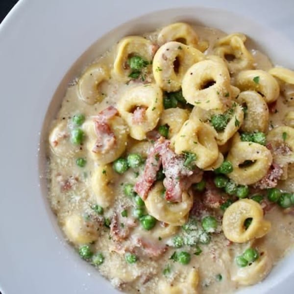 Tortellini pasta in a creamy sauce with peas, bacon, and parsley in a white bowl.