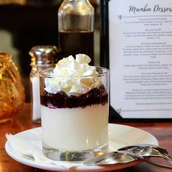 A dessert glass with white pudding, berry compote, and whipped cream on a wooden table.