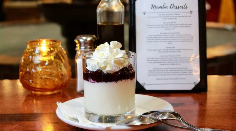 A dessert glass with white pudding, berry compote, and whipped cream on a wooden table.