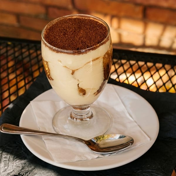 Tiramisu dessert in a glass with a spoon on a white plate, on a metal table.