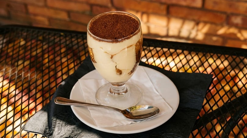 Tiramisu dessert in a glass with a spoon on a white plate, on a metal table.