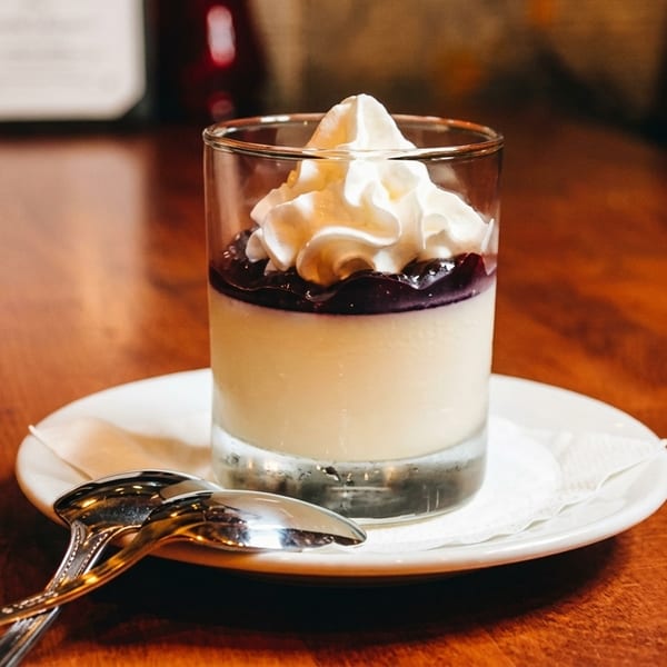 A glass of panna cotta with blueberry topping and whipped cream on a wooden table.