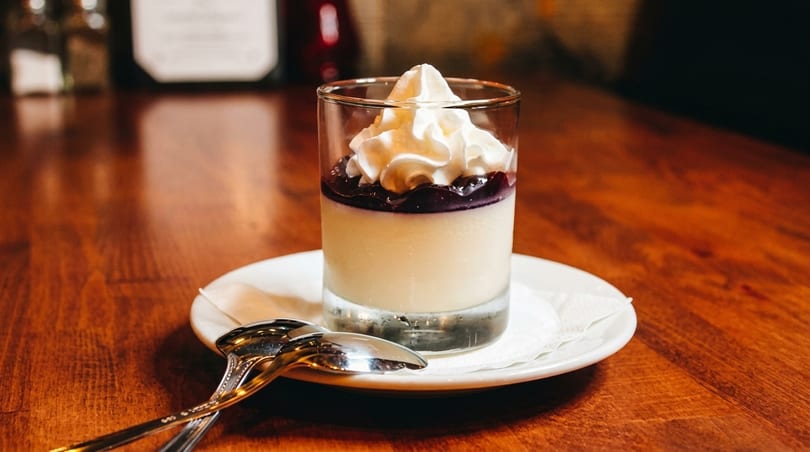 A glass of panna cotta with blueberry topping and whipped cream on a wooden table.
