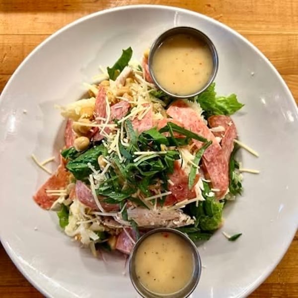 A large salad with greens, sliced salami, shredded cheese, and two cups of creamy dressing.