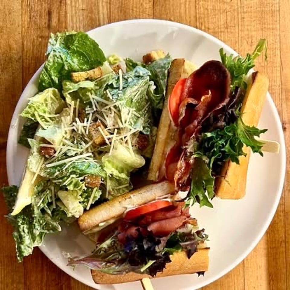 Plate of baguette sandwiches with leafy greens, tomatoes, cured meat, and a side Caesar salad on a wooden table.